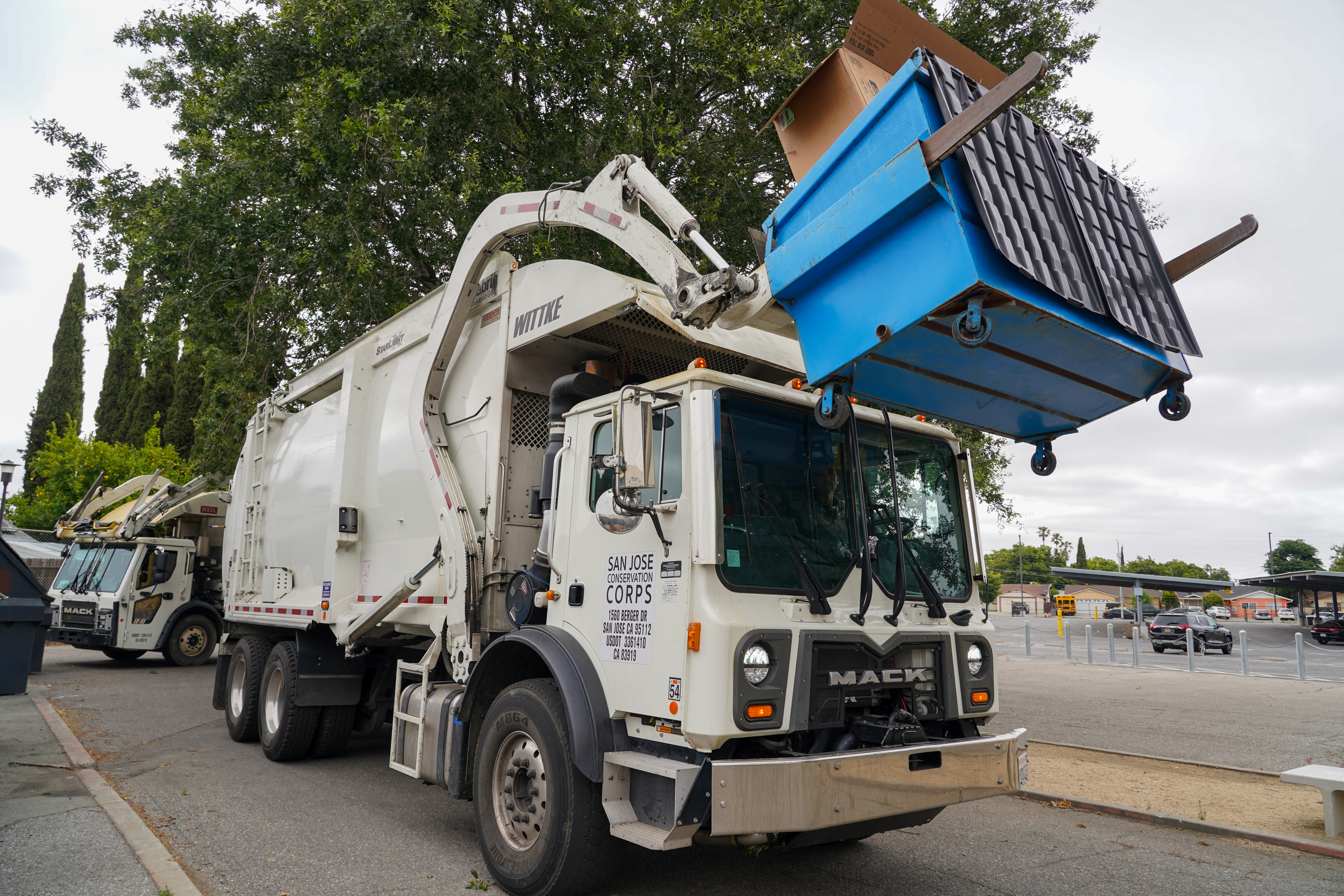 Garbage truck lifting blue dumpster – A white garbage truck is using its front-loading arms to lift a blue dumpster filled with cardboard.