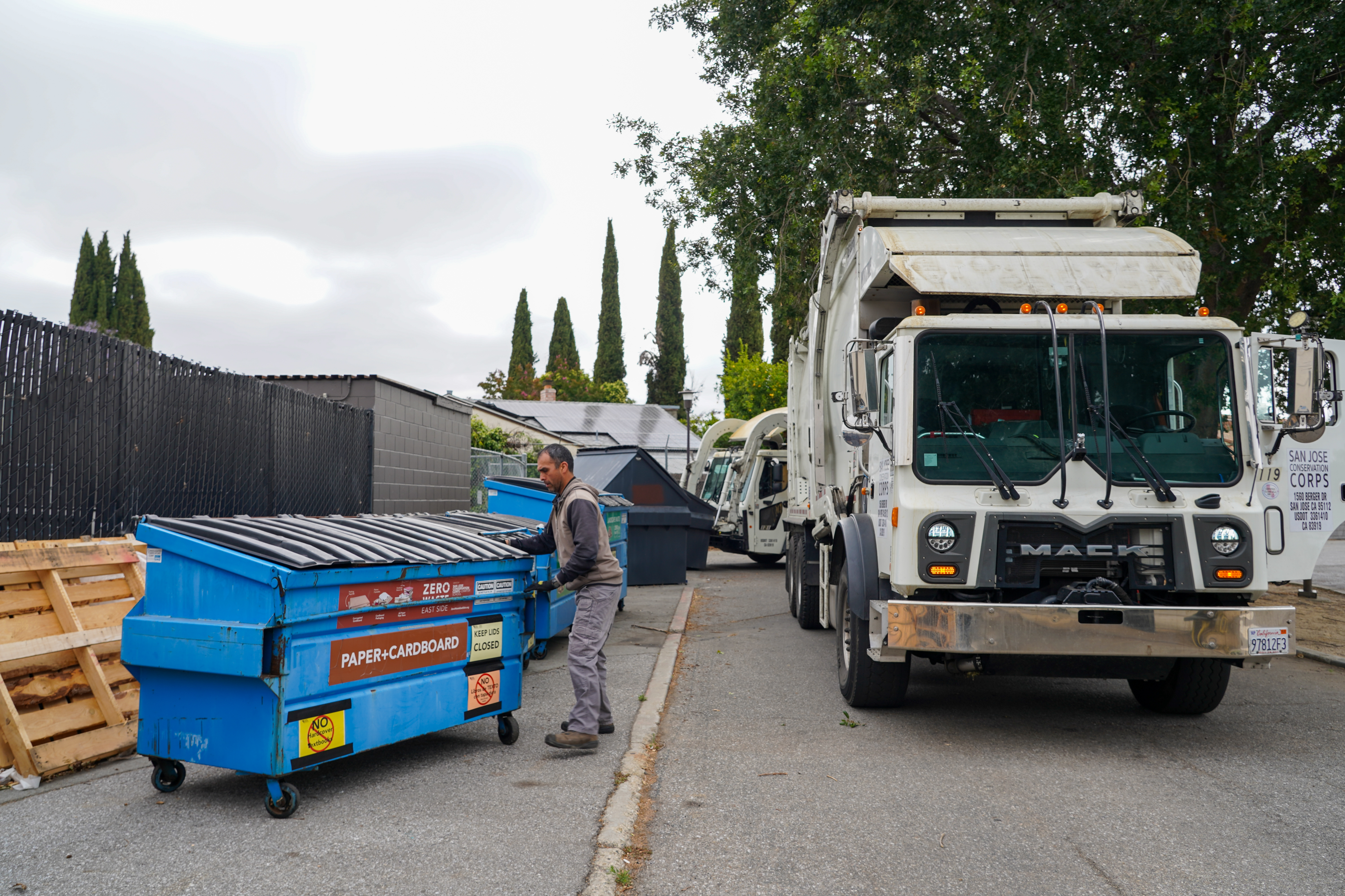 Corp member moving recycling dumpster – A corp member pushes a blue recycling dumpster labeled “Paper + Cardboard” near a garbage truck.
