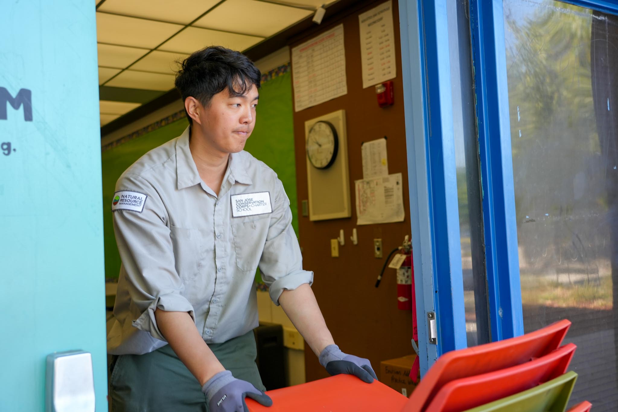 Corp member carrying red chairs – A corp member wearing gloves carries stacked red chairs out of a classroom.