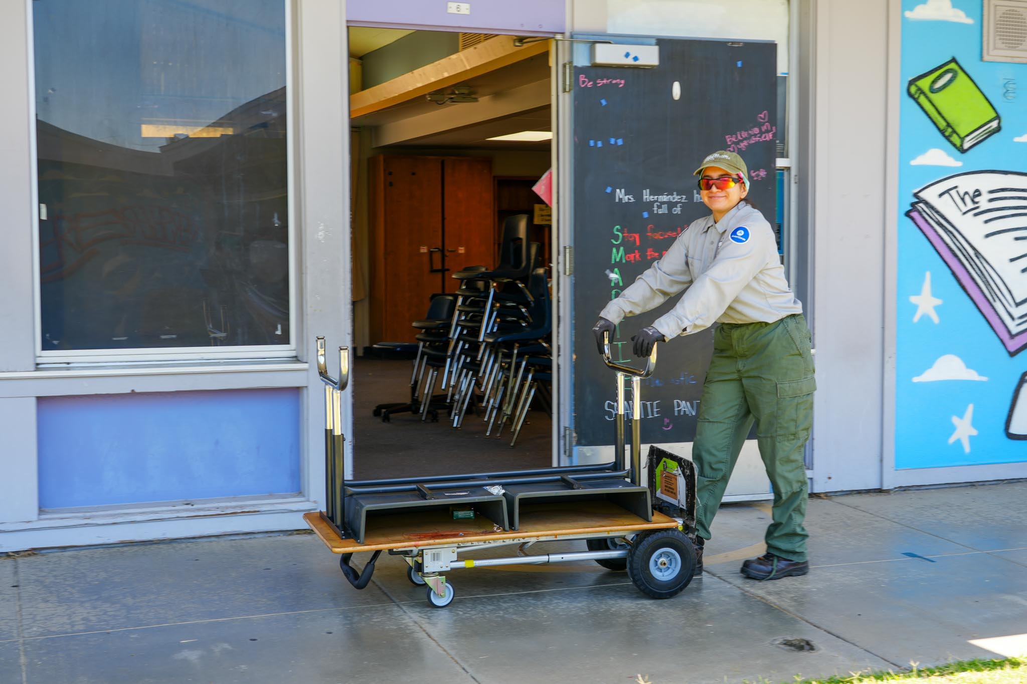 Corp member moving tables on a cart – A corp member pushes a flat cart loaded with upside-down tables out of a classroom doorway.