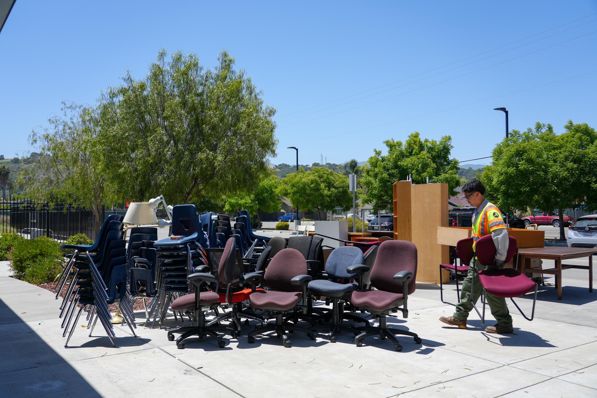 Corp member moving office chairs outdoors – A corp member carries a maroon office chair while standing near piles of stacked chairs and desks outside.