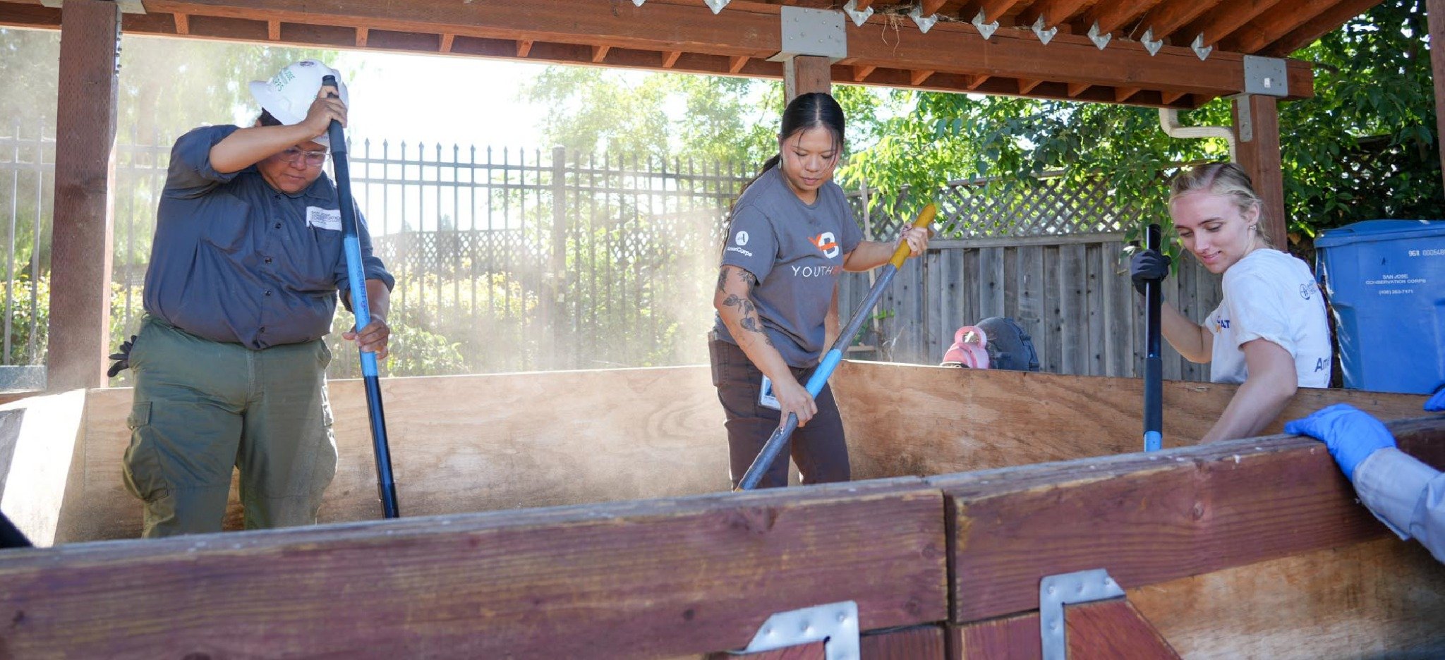 Corp members composting vegetables – Corp members under a wooden shelter prepare and add leafy vegetables to a large compost bin.