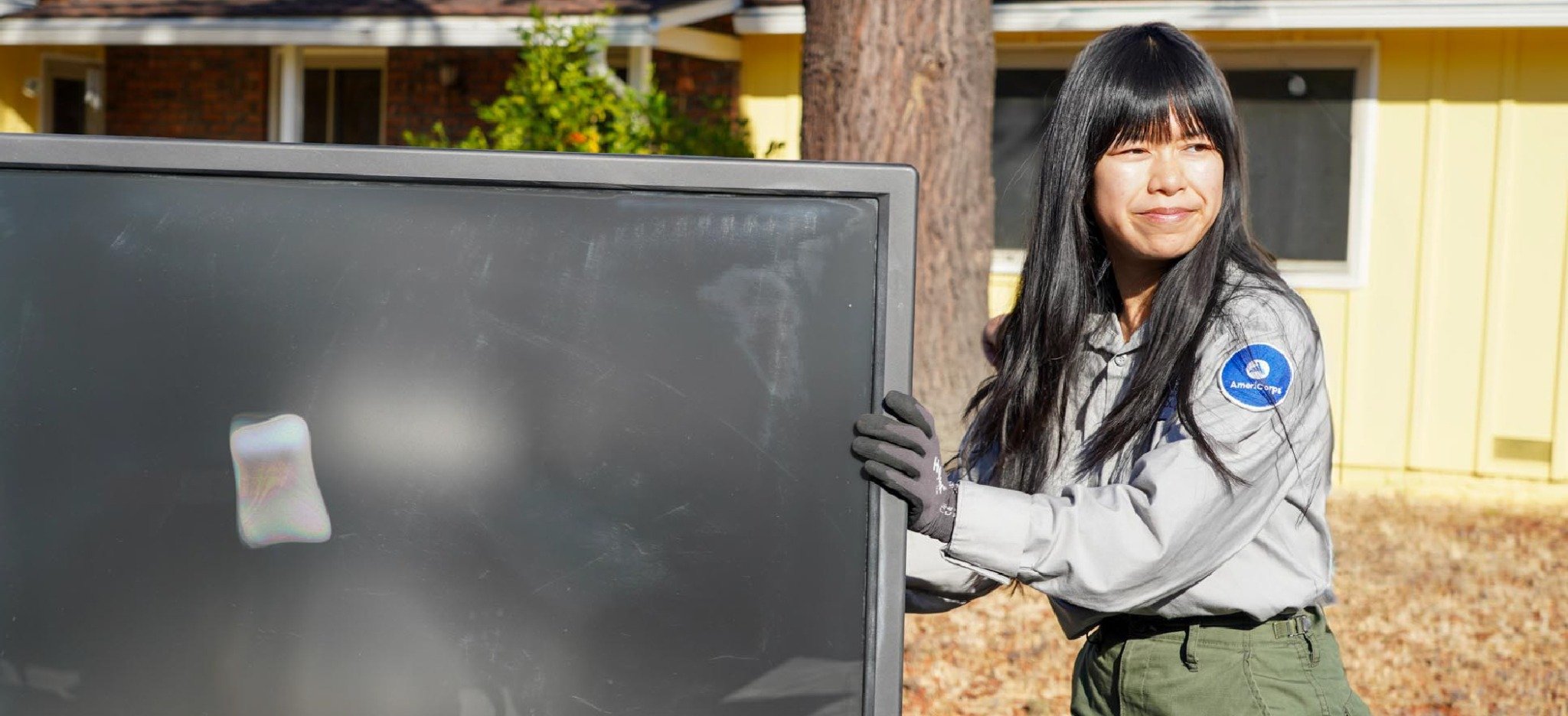 Corp member collecting e-waste – A corp member wearing gloves lifts and carries a large television outdoors as part of an electronic waste recycling program.